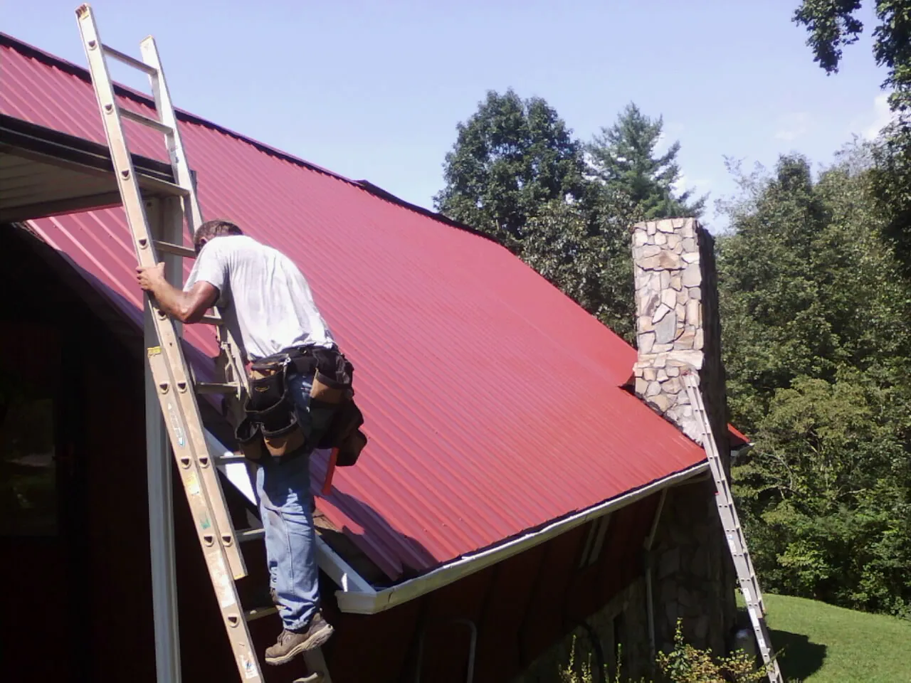 Roofer on a ladder beside a red metal roof and stone chimney — Soffit & Fascia Repair by Roofing Brothers in Lexington